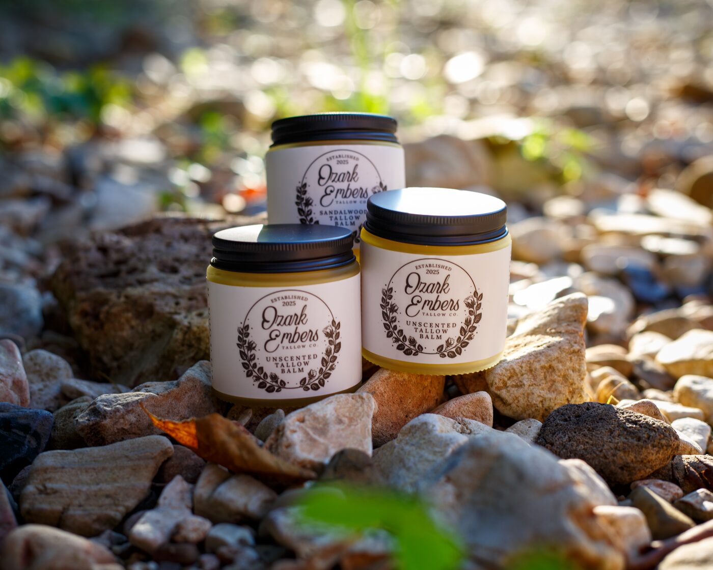 Two jars of unscented tallow balm and one of sandalwood, stacked on creek stones beside a fallen leaf